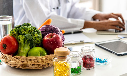 Close up image of a tomato, broccoli, apple, cabbage and carrot with a pair of hands typing on a laptop in the background