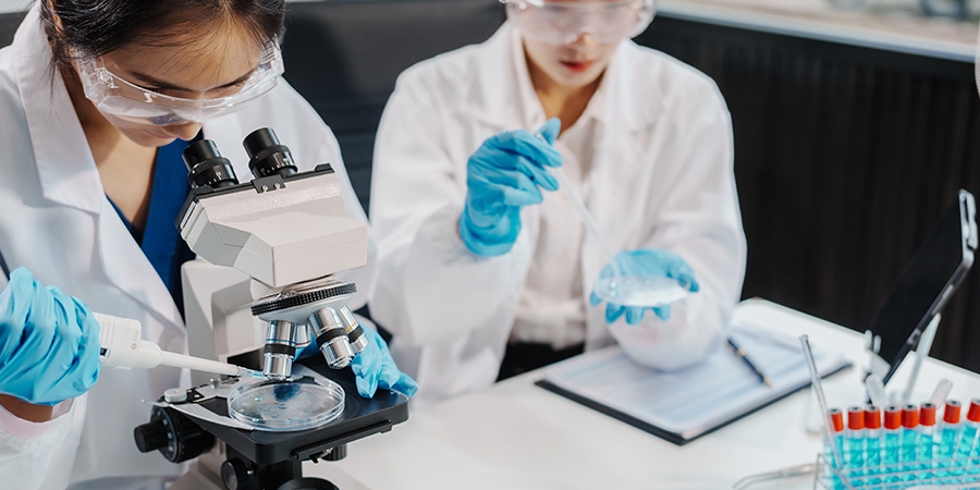 Two female researchers work at a desk in a lab, analyzing liquid samples under a microscope, experimenting with chemistry, and using high technology for important scientific discoveries.