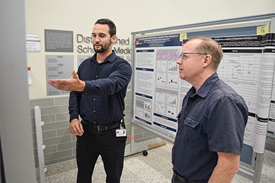 Two men are facing a mounted poster, one is holding his arm out to show the other something on the poster.